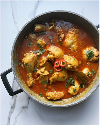 overhead-shot-open-pot-with-one-black-handle-showing-on-marble-counter-with-chcken-curry-inside-in-red-sauce-and-green-herbs-dotted-on-top